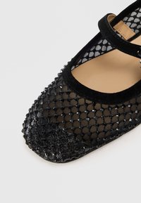 Chaussure plate noire avec un dessus en maille présentant un motif. L'orteil est orné d'un accent texturé perlé et d'une sangle en daim sur le coup de pied.