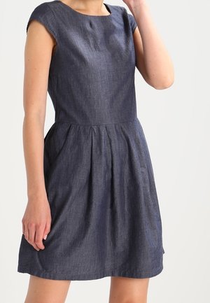 Kvinde iført en mørkeblå chambray-kjole med korte ærmer, knælængde, tætsiddende overdel og plisseret nederdel.