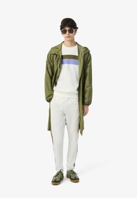 Chaqueta ligera color verde oliva, sudadera blanca con rayas horizontales en verde oliva, azul y púrpura, pantalones blancos y zapatillas verdes con suelas marrones.
