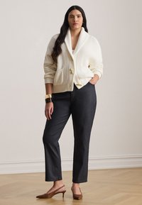 Γυναίκα με μακριά σκούρα μαλλιά, φοράει λευκή πλεκτή ζακέτα, μαύρο παντελόνι, καφέ πέδιλα με λουράκι και χρυσά και σκούρα βραχιόλια, στέκεται σε ξύλινο πάτωμα.