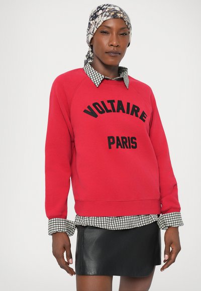 Rode sweatshirt met zwarte tekst "VOLTAIRE PARIS", gelaagd over een zwart-witte geblokte blouse. De outfit is gecombineerd met een zwarte leren rok.