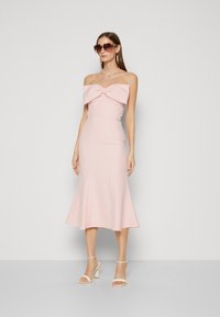 Abito midi rosa senza spalline con un grande fiocco dettagliato sulla scollatura e un orlo svasato. Il modello indossa scarpe con tacco nude e occhiali da sole oversize.