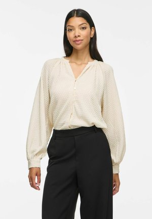 Vrouw draagt een gestructureerde crèmekleurige blouse met lange pofmouwen en zwarte hooggetailleerde broek, staand tegen een witte achtergrond.
