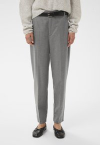 Persona che indossa pantaloni grigio a righe con una cintura nera, un maglione beige e ballerine nere, in piedi su uno sfondo bianco.