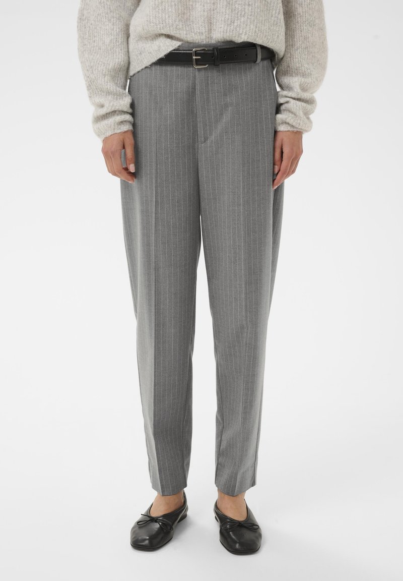 Persona che indossa pantaloni grigio a righe con una cintura nera, un maglione beige e ballerine nere, in piedi su uno sfondo bianco.