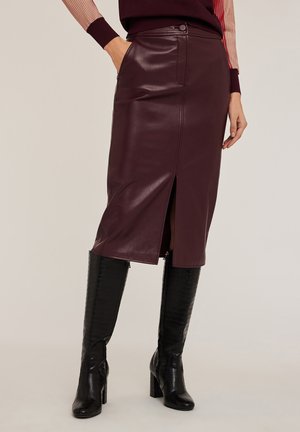 Jupe midi en simili cuir bordeaux avec fente avant, au design ajusté et deux poches latérales. Associée à des bottes noires à talons hauts, montant jusqu'aux genoux.