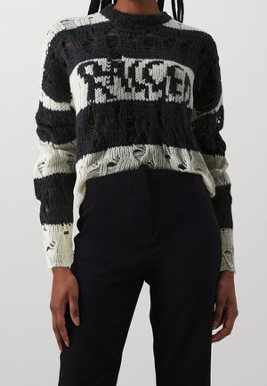 Pull en tricot noir et blanc usé avec des trous et un texte blanc audacieux sur la poitrine, associé à un pantalon noir taille haute.