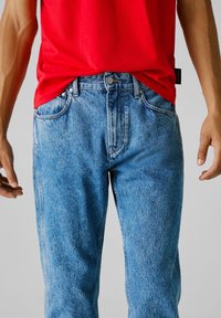 Hombre con camisa roja y jeans de mezclilla azules, con las manos relajadas a los lados frente a un fondo gris liso.