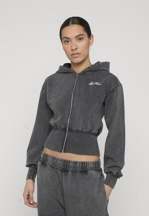 Kobieta ubrana w czarną, spraną, zapinaną na zamek krótką bluzę z kapturem oraz dopasowane spodnie dresowe z elastycznym pasem na jednolitym tle.