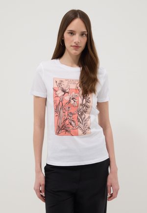 Giovane donna con capelli lunghi castani indossa una maglietta bianca con schizzo floreale rosa e testo, abbinata a pantaloni neri, in piedi davanti a uno sfondo semplice.