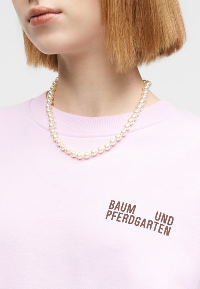 Ανοιχτό ροζ φούτερ με στρογγυλή λαιμόκοψη, διακοσμημένο με το κείμενο «BAUM UND PFERDGARTEN» σε σκούρο καφέ, συνοδευόμενο από χάντρες ψεύτικων μαργαριταριών.