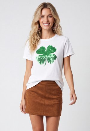 Donna sorridente con lunghi capelli biondi indossa una maglietta bianca con un grande trifoglio verde e una minigonna in pelle scamosciata marrone su uno sfondo semplice.