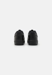 Black sneakers with a textured material, rounded back, and a logo detail on the heel. Features a flat sole and reinforced stitching.