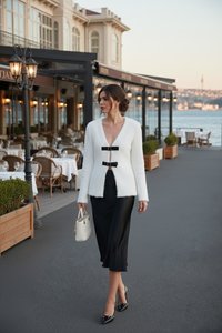 Woman in white cardigan and black skirt walking along waterfront restaurant patio holding a white handbag at dusk.