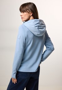 Světle modrá mikina na zip s měkkou texturou, opatřená kapucí a logem na levém rukávu, kombinovaná s tmavými džíny.