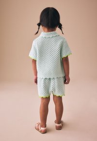Niño con cabello trenzado que está de pie, vistiendo una camiseta de manga corta texturizada azul claro y pantalones cortos a juego con ribete amarillo, y sandalias sobre fondo beige.