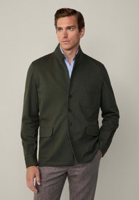Giacca leggera di un verde scuro, con colletto alto, bottoni frontali e due tasche applicate. Indossata sopra una camicia azzurro chiaro. Tessuto testurizzato.