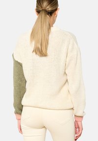 Kvinna med blont hästsvans som bär en krämfärgad stickad tröja med en olivgrön vänsterärm och rosa manschetter, i kombination med krämfärgade byxor, vänd bort.