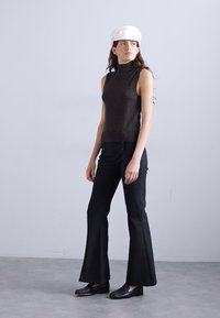 Top senza maniche color marrone con collo alto abbinato a pantaloni neri a palazzo e mocassini neri. La modella indossa un cappellino bianco, in piedi su uno sfondo semplice.