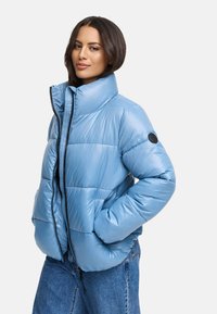 Light blue puffer jacket with a high collar, zip closure, and elastic cuffs. Features a circular logo on the left sleeve. Smooth, shiny texture.