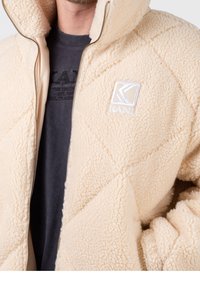 Beige zip-up jacka med en quiltad textur och fleece-material. Har en logotyp på bröstet och ett innertyg i en mörkare nyans.
