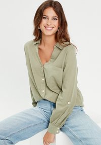 Chemise vert olive à boutons avec un col en V, poche poitrine et manches longues à revers. Associée à un jean taille haute bleu clair.