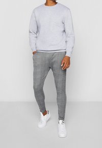 Mężczyzna stoi z rękami w kieszeniach, ubrany w jasnoszary sweter, szare spodnie w kratę o dopasowanym kroju i białe sneakersy na jednolitym tle.