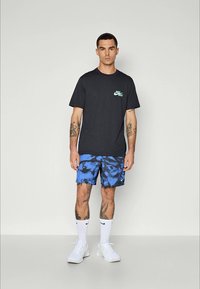 Sort bomulds-T-shirt med grønt logo på brystet, parret med blå tie-dye shorts. Hvide sneakers og sokker fuldender det sporty outfit.