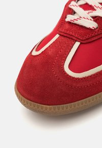 Baskets rouges avec une tige en daim et des accents en nylon, détails crème, semelle en caoutchouc gomme texturée et design de lacets classique.