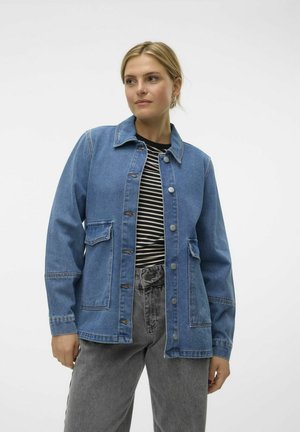 Veste en denim bleu moyen avec un devant à boutons, deux poches avant et un col classique. Portée par-dessus une chemise à rayures noir et blanc.