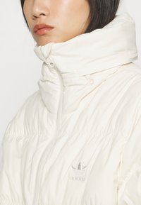 Veste matelassée couleur crème avec un col montant, texture côtelée et un logo argenté sur la poitrine. Dispose d'un cordon de serrage ajustable au niveau du cou.