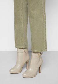 Beżowe botki na obcasie z teksturowanym wykończeniem, z zamkiem błyskawicznym z boku i regulowanym paskiem. Ustawione na tle jasnoszarych spodni.