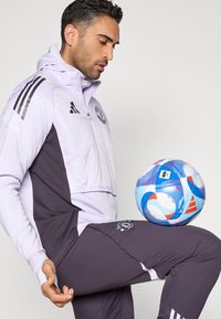 Lys lilla og mørkegrå sportjakke med glidelås og logo, kombinert med matchende bukser. En flerfarget fotball balanseres på et opphøyd kne.