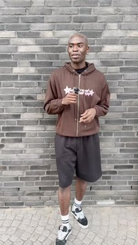 Brun hettejakke med hvite og rosa stjernetegninger, svarte shorts og svarte joggesko med hvite detaljer, foran en grå mursteinsbakgrunn.