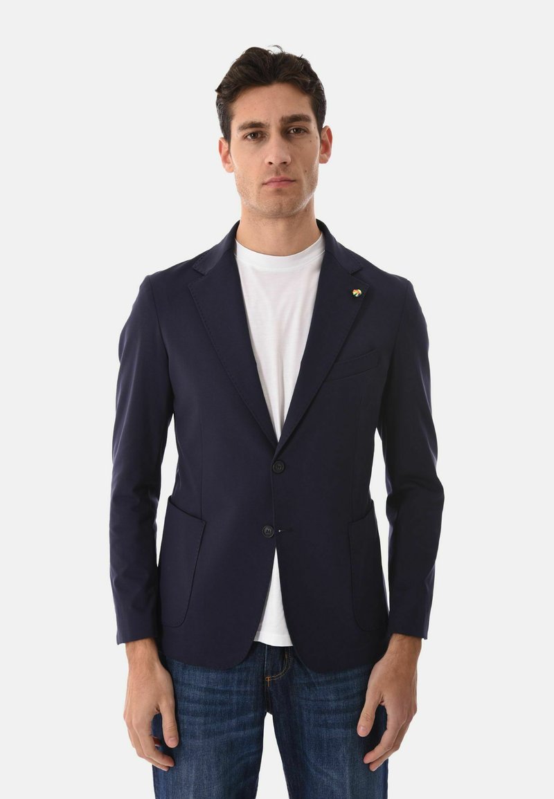 Giovane uomo che indossa un blazer blu navy sopra una t-shirt bianca e jeans blu, in piedi contro uno sfondo chiaro e uniforme.