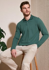 Camisa polo de manga larga verde hecha de tela suave, con cuello y un brillo sutil, combinada con pantalones slim-fit de un beige claro.