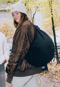 Hnědá bombardovací bunda s žebrovaným lemem, černá taška přes rameno, bílá pletená čepice a světlé kalhoty, v přírodním prostředí s podzimním listím.