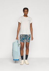 Φωτεινή μπεζ μπλούζα, σορτς με σχέδιο μπλε και πράσινα φύλλα, κρατώντας ανοιχτό μπλε σακίδιο. Κρεμ παπούτσια, μαύρες κάλτσες. Στάση όρθια.