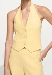 Žlutá halenková vesta na zavazování s hlubokým výstřihem do V, třemi knoflíky a dvěma předními kapsami; hladká textura a přiléhavý střih.
