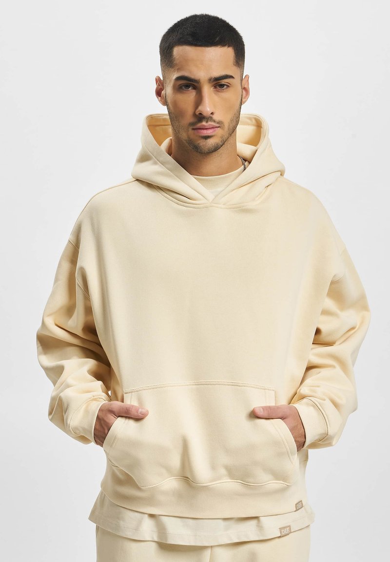 Beige oversized hættetrøje lavet af blødt stof med en stor forlomme og hætte med snøring. Den nederste kant inkluderer et mærke.