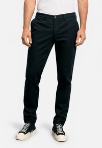 Pantaloni chinos neri in misto cotone, con taglio dritto, chiusura con bottoni e tasche laterali, abbinati a sneakers scure.