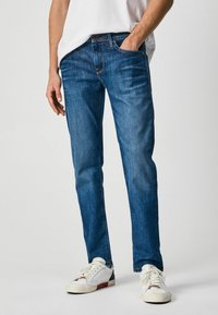 Jeans de denim azul oscuro con un ajuste slim, bolsillos abiertos y un diseño clásico de cinco bolsillos, combinados con zapatillas blancas.