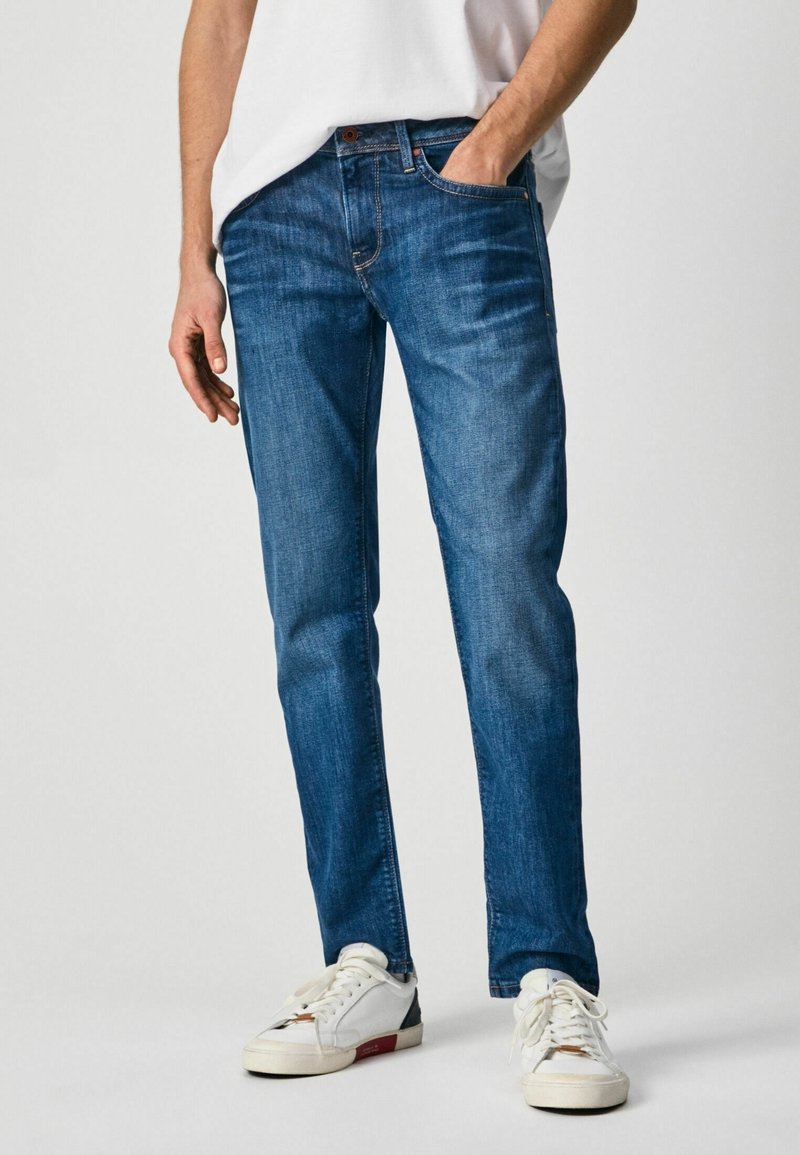 Jeans de denim azul oscuro con un ajuste slim, bolsillos abiertos y un diseño clásico de cinco bolsillos, combinados con zapatillas blancas.