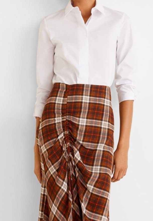 Biała koszula z kołnierzykiem w komplecie z brązowo-pomarańczową spódnicą w kratkę, z zaszewkami po bokach i gładką teksturą materiału.