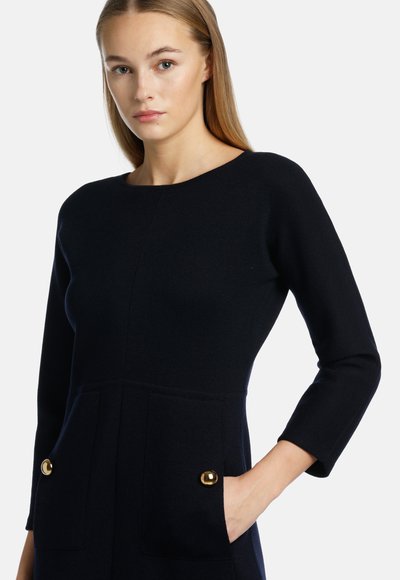 Strickkleid in Marineblau mit rundem Ausschnitt, dreiviertel Ärmeln, zwei vorderen Taschen mit goldenen Knopfakzenten und einer taillierten Silhouette.