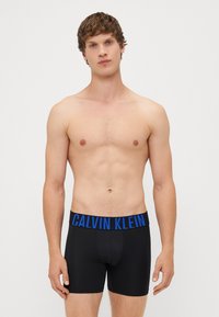 Boxeri negri din bumbac, cu un brâu logo Calvin Klein albastru, design ajustat și lungime până la mijlocul coapsei, prezentati pe un model masculin.