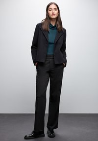 Schwarzer Blazer über einem türkisfarbenen Top mit Rollkragen, kombiniert mit dunkelgrauen, maßgeschneiderten Hosen. Glänzende schwarze Schuhe runden das Outfit ab.