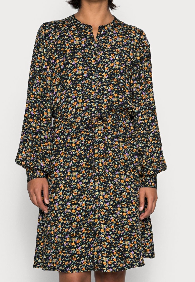 Femme portant une robe noire à manches longues, longueur genou, avec un petit imprimé floral multicolore et des boutons sur le devant.