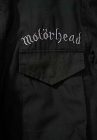Černá látková bunda s výrazným šedým vyšívaným logem "Motörhead" nad klopou kapsy s špičatým tvarem.