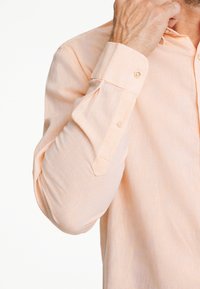 Camisa de manga larga en tejido de color durazno claro con acabado texturizado, que presenta un puño y un collar abotonados. Variaciones sutiles en el color.